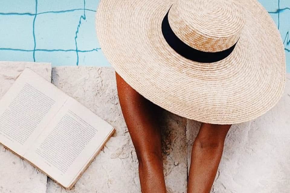 Person poolside wearing oversized straw hat, reading book by turquoise water