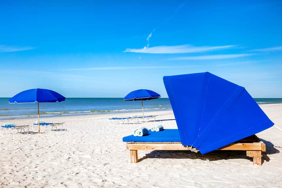 Sandy beach with blue umbrellas, lounge chairs, and clear sky with calm ocean in background