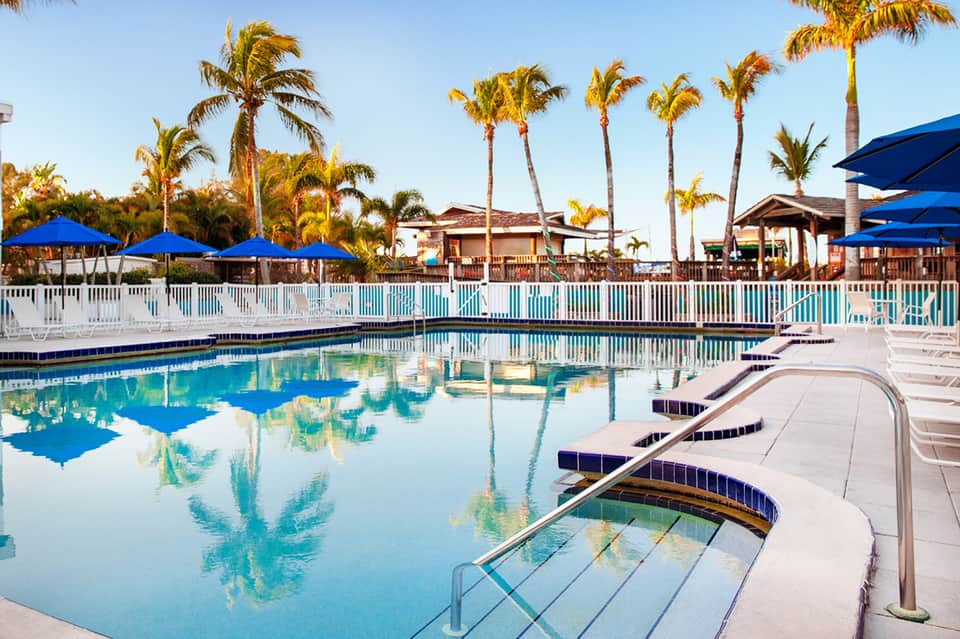 Resort pool with palm trees, blue umbrellas, white fence, and tropical clubhouse architecture