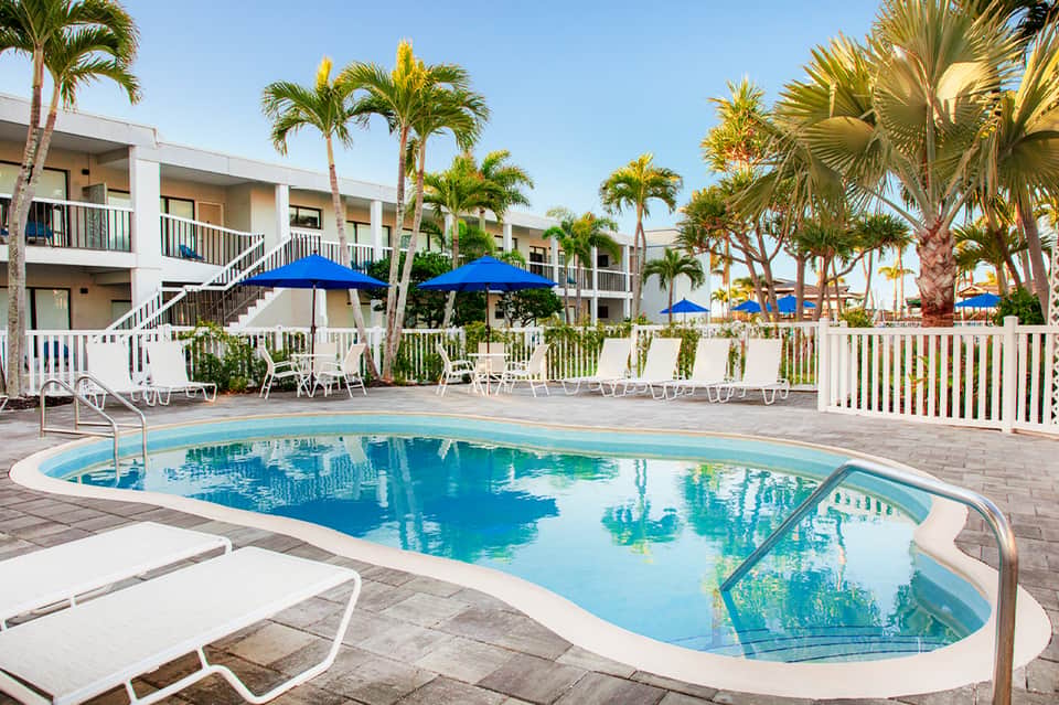 Resort pool area with blue umbrellas, lounge chairs, palm trees, and white residential buildings