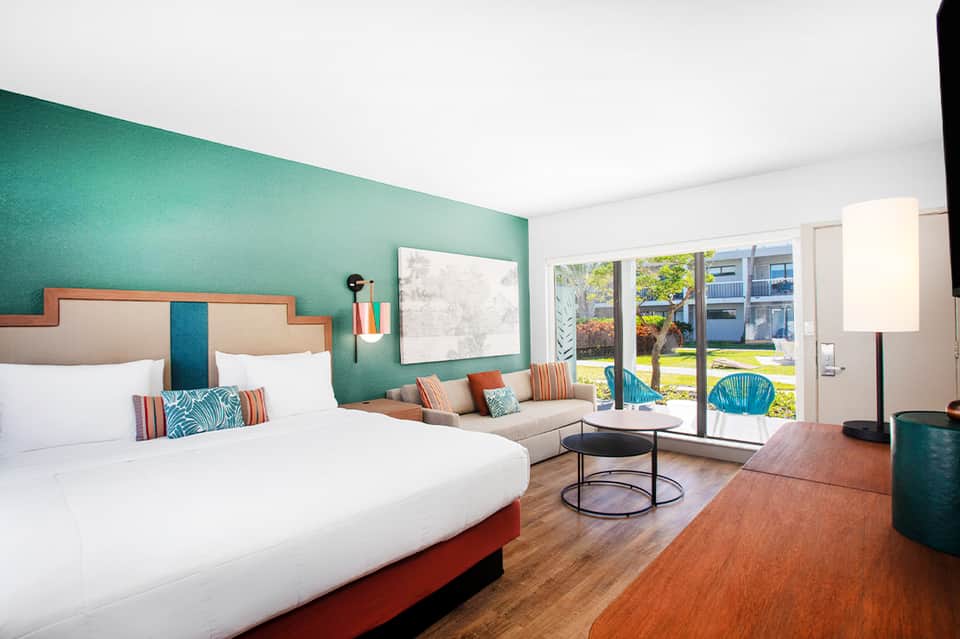 Modern hotel room with turquoise accent wall, white bedding, seating area, and patio doors