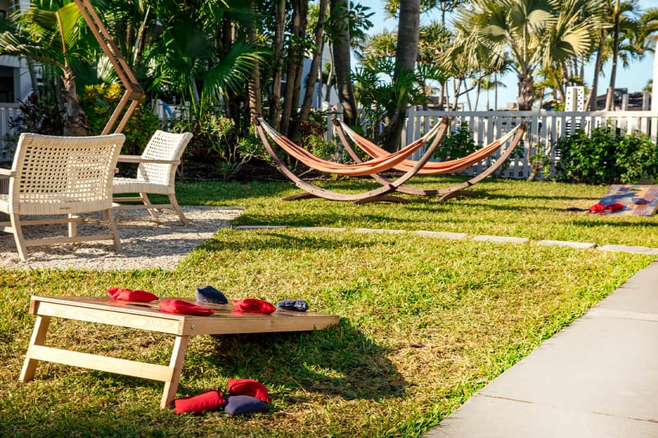 Beach lawn games area with cornhole boards, hammocks, and lounge chairs under palm trees