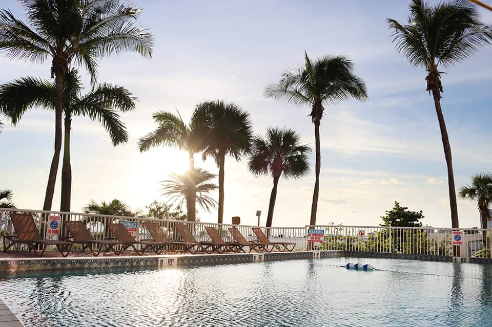 Resort swimming pool with palm trees reflected in still water and beachfront buildings in background