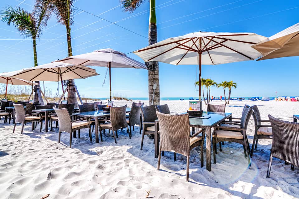 Beachfront dining area with umbrellas, wicker chairs, and ocean view under clear blue sky