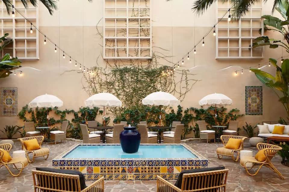 Courtyard pool area with white umbrellas, woven lounge chairs, ivy-covered walls, and string lights overhead