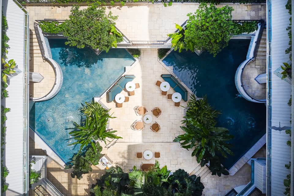 Overhead view of two curved pools with lounge chairs and tropical vegetation between buildings