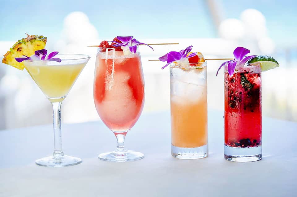Four colorful cocktails with tropical garnishes and purple flowers against a sky background