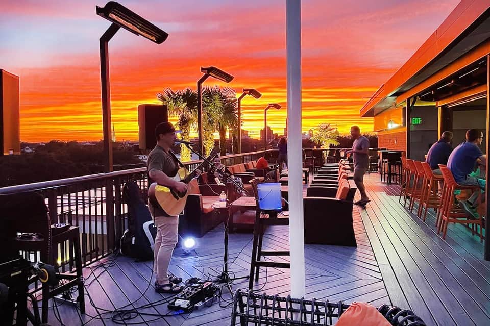 Live musician performing on a rooftop terrace at golden sunset with palm trees and city skyline behind