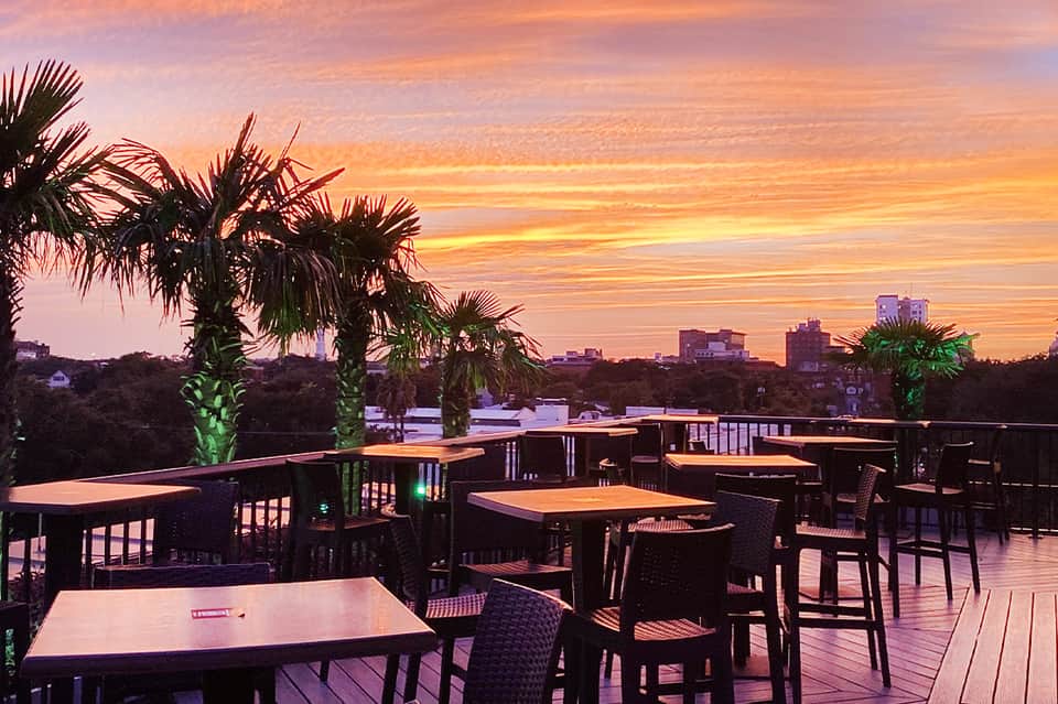 Outdoor rooftop dining area at sunset with palm trees, wooden tables, and colorful sky reflection