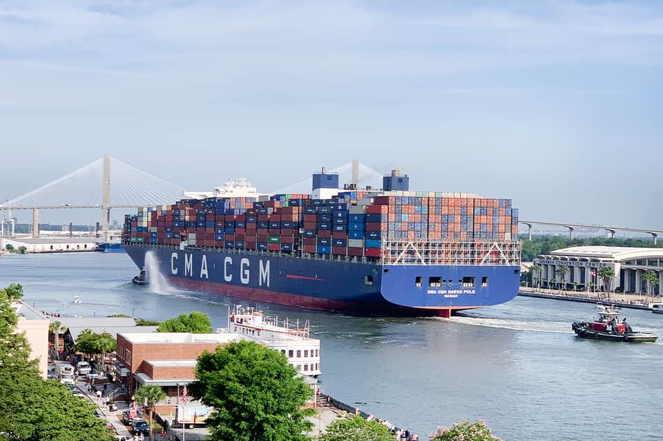 Large cargo ship with stacked shipping containers passing under bridge near waterfront city