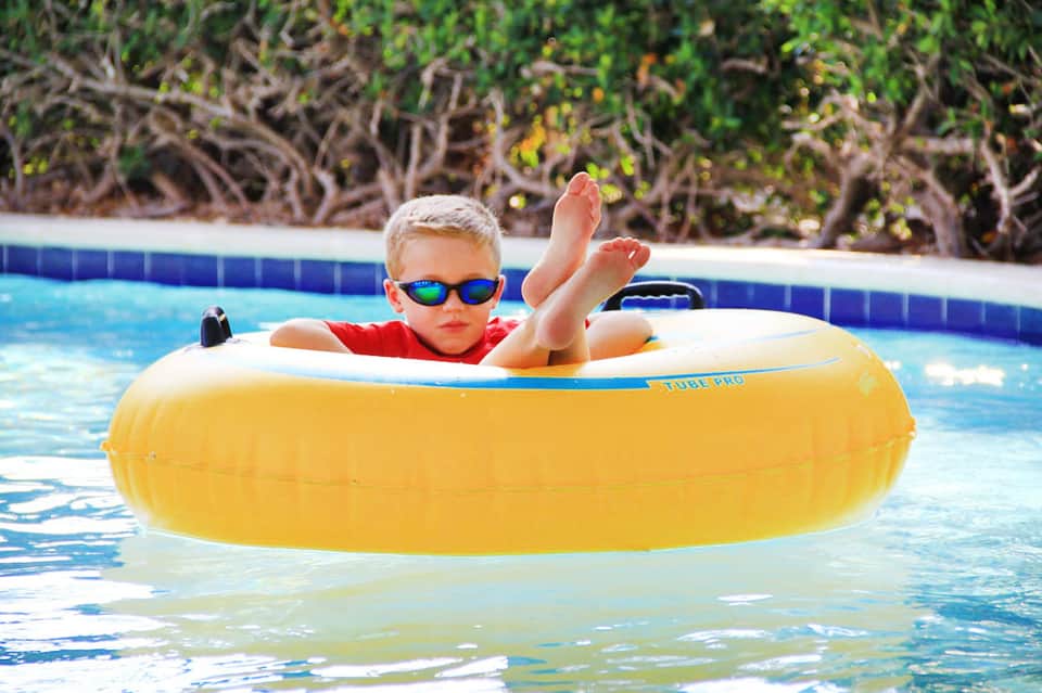 Young child in red shirt and blue sunglasses floating in yellow tube in pool with landscaped background