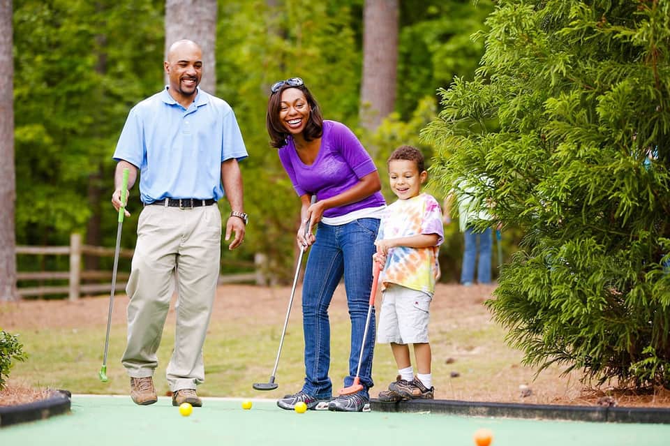 Family of three playing mini golf outdoors surrounded by green landscaping and trees