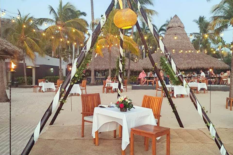 Beach bonfire dinner setup with tiki torches, white tablecloth, and palm surroundings