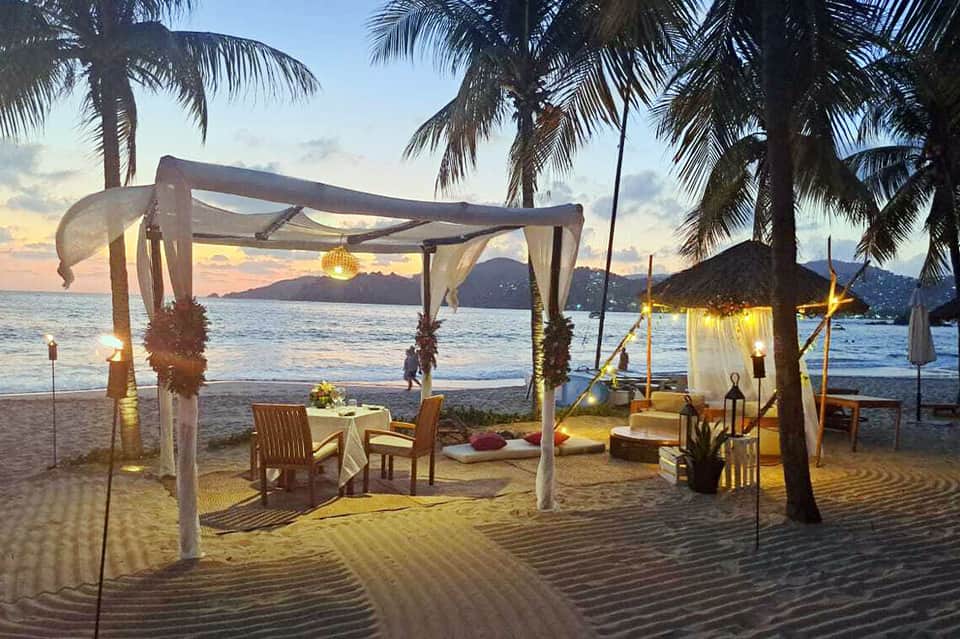 Beachfront dinner setup with white canopy, tables, and palm trees at sunset with water views