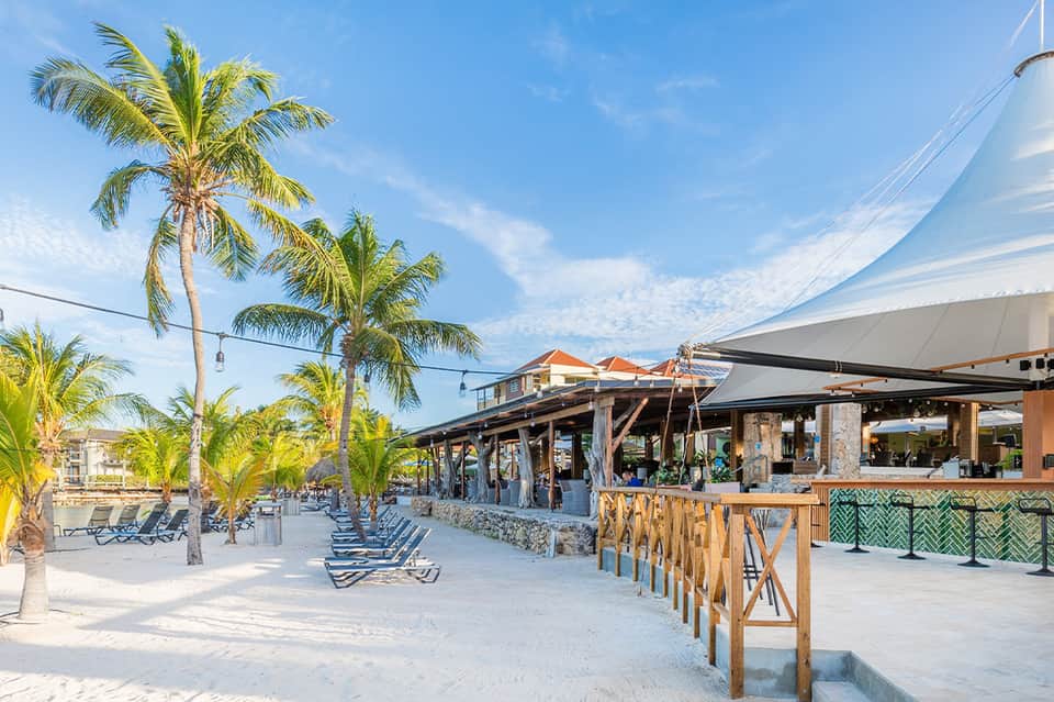 Beachfront restaurant and bar with retractable shade sails, palm trees, lounge chairs, and sandy beach seating