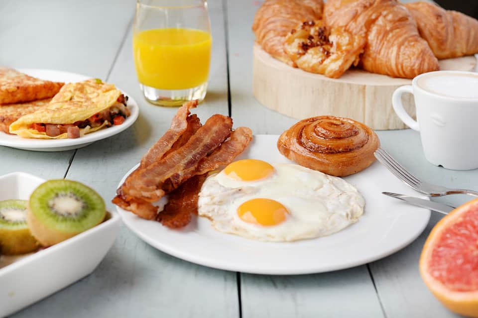 Breakfast spread with eggs, bacon, croissants, fresh fruit, juice, and pastries on white table