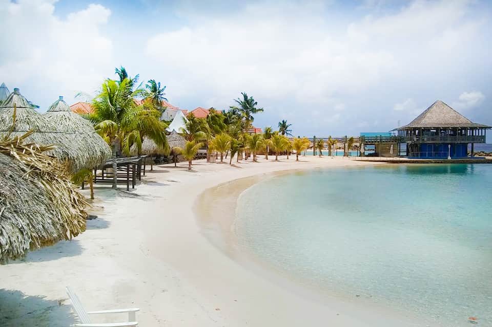 Tropical beach resort with white sand, turquoise water, thatched umbrellas, palm trees, and beachfront buildings