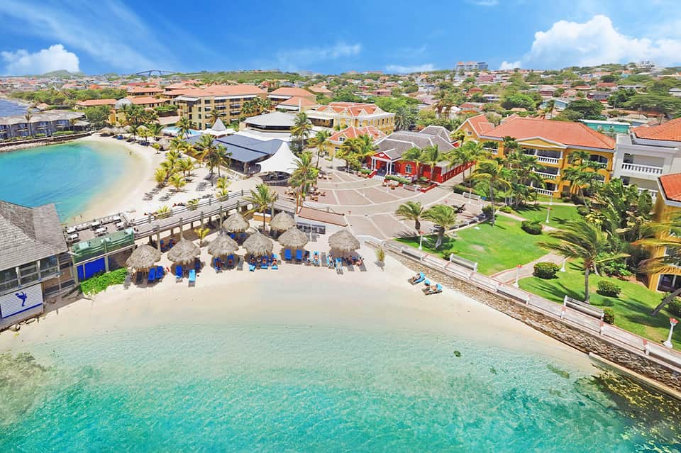 Aerial view of colorful beachfront resort with turquoise water, umbrellas, and multi-story buildings