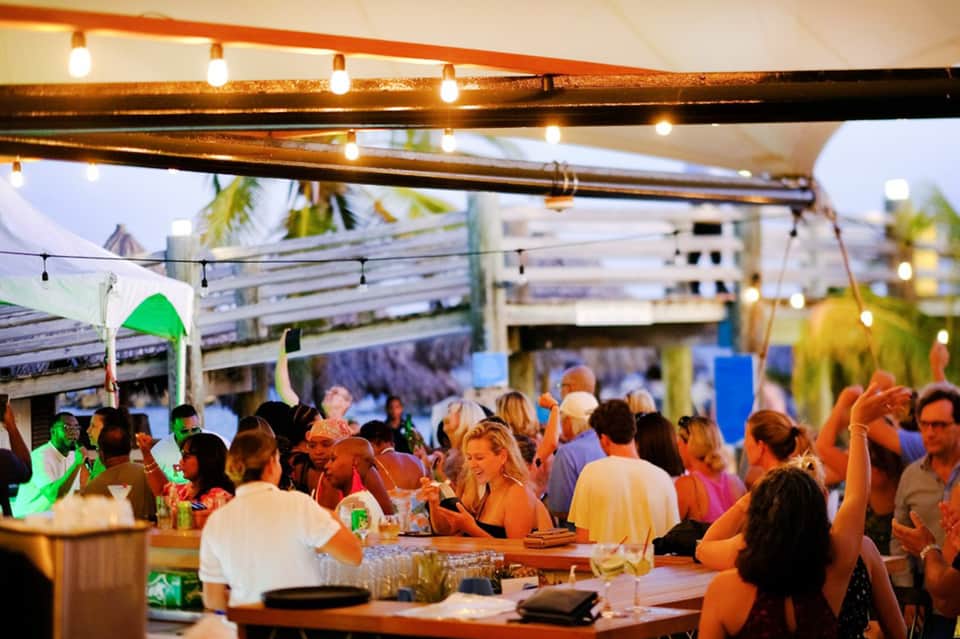 Crowded beach bar event at sunset with string lights and palm tree decorations