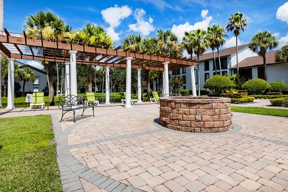 Outdoor courtyard with pergola structure, fire pit, palm trees, manicured lawn, and resort building architecture.