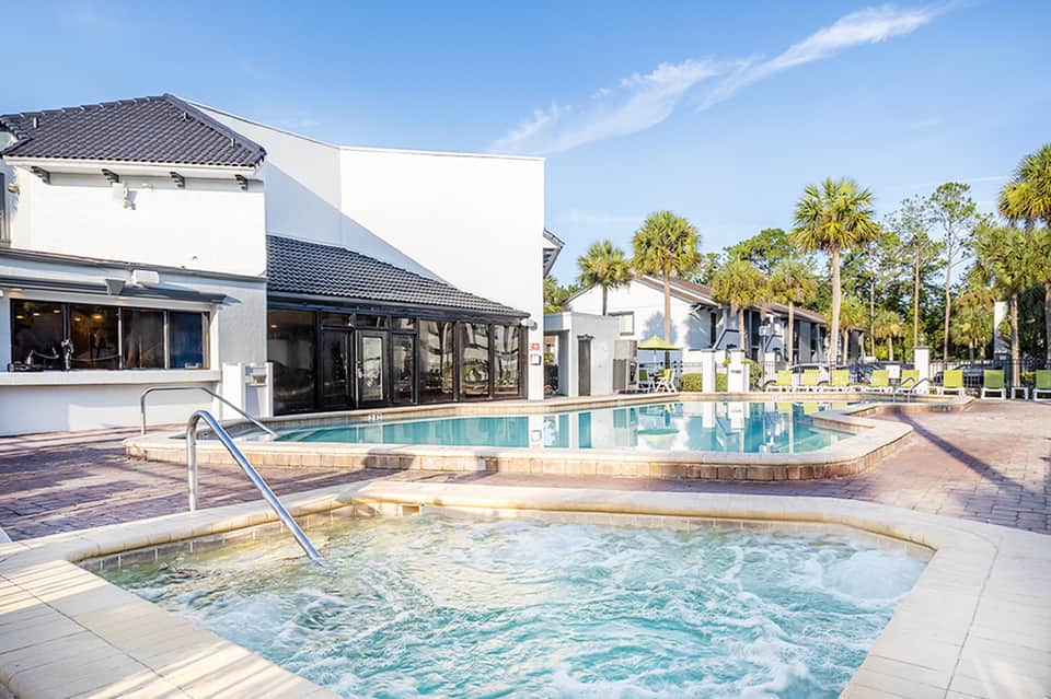 Resort pool and hot tub area with white buildings, palm trees, and poolside cabanas