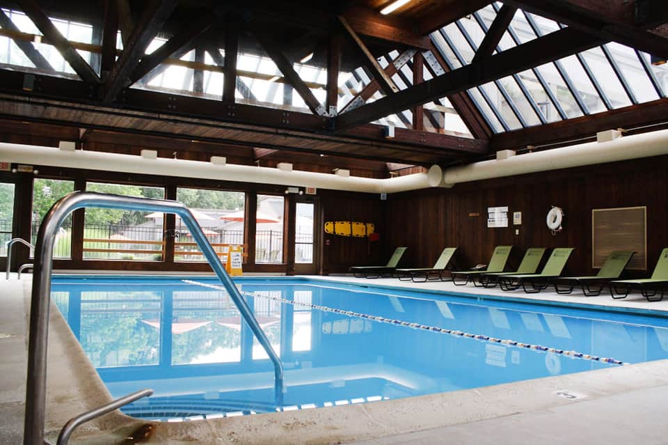 Indoor lap pool with blue water, skylights, wooden ceiling beams, and green lounge chairs along the side