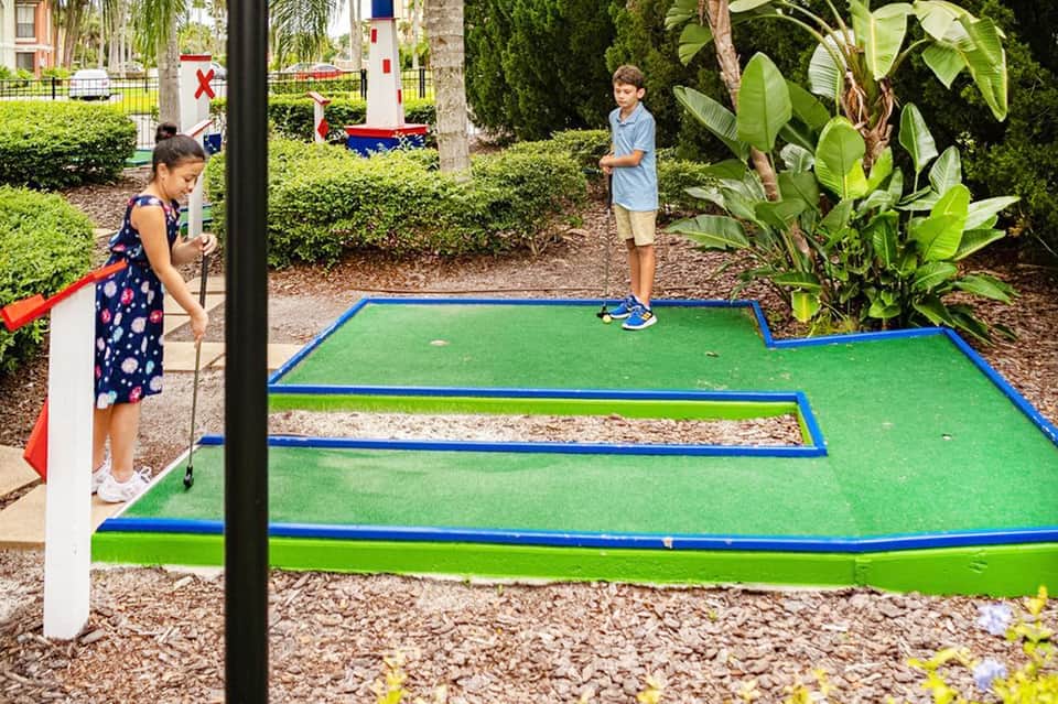 Children playing mini golf outdoors on green course with tropical landscaping