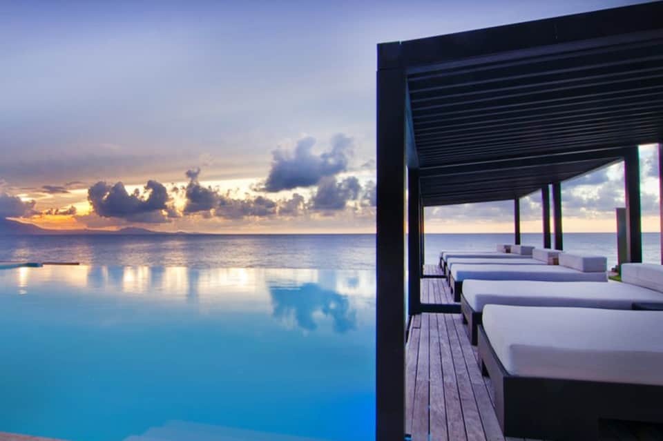 Infinity pool overlooking ocean at sunset with modern white lounge chairs and wooden deck