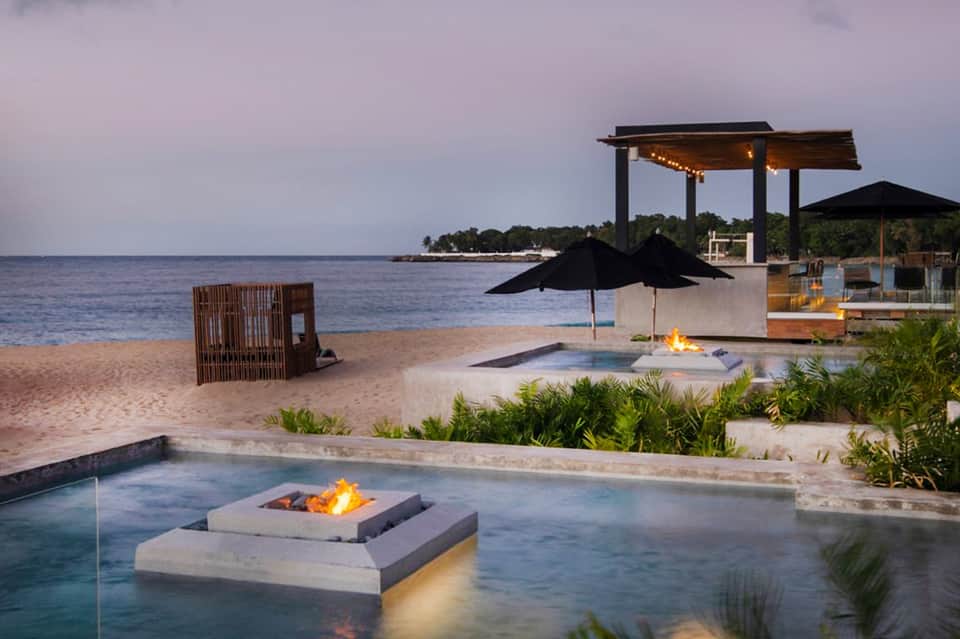 Beachfront fire pit lounge at dusk with modern pergola, fire feature, and ocean views with resort buildings.