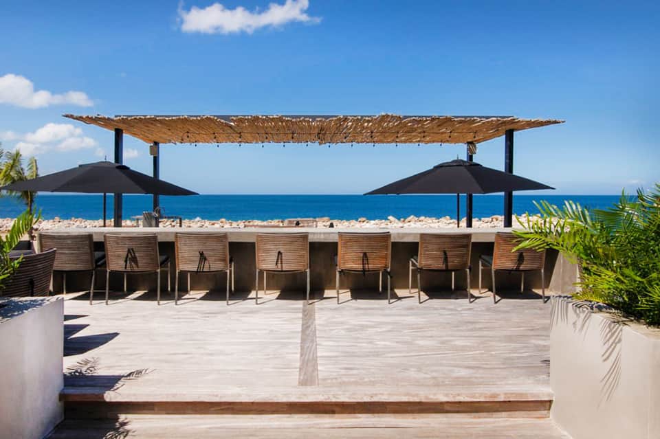 Beachfront dining area under woven roof with umbrellas overlooking turquoise ocean