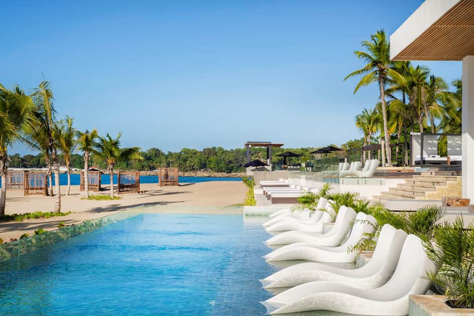 Modern resort pool with white curved lounge chairs, palm trees, and beachfront access