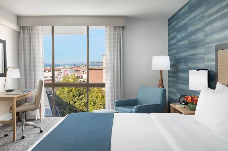 Hotel bedroom with ocean view windows, blue accent chair, desk area, and modern coastal décor