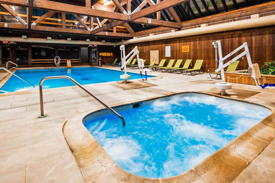 Indoor pool area with hot tub, wooden beams, lounge chairs, and white accessibility lifts