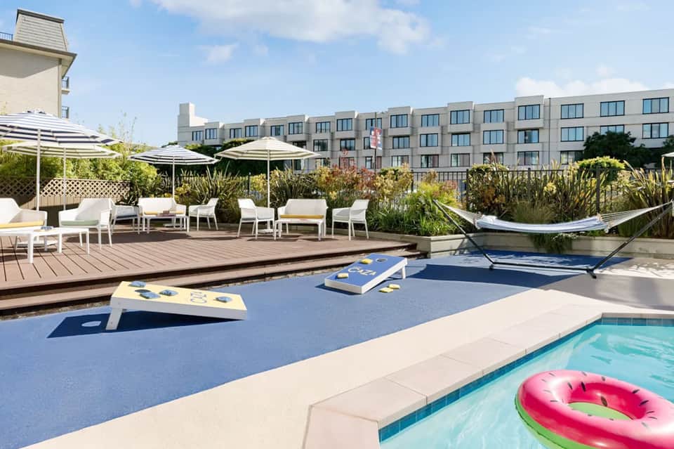 Resort pool deck with cornhole games, lounge chairs, umbrellas, and modern apartment buildings