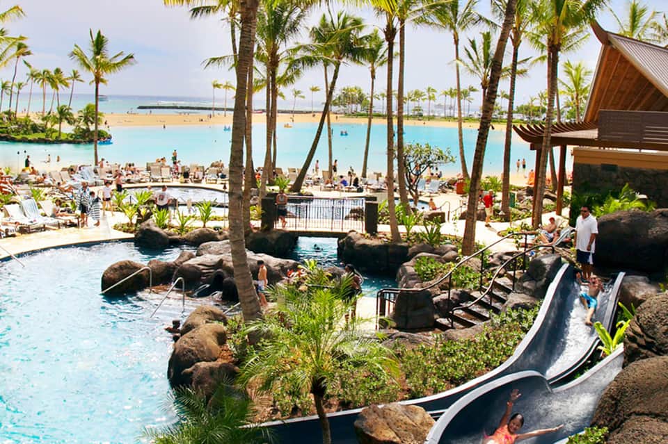 Tropical resort pool area with turquoise water, sand beach, palm trees, and water slide
