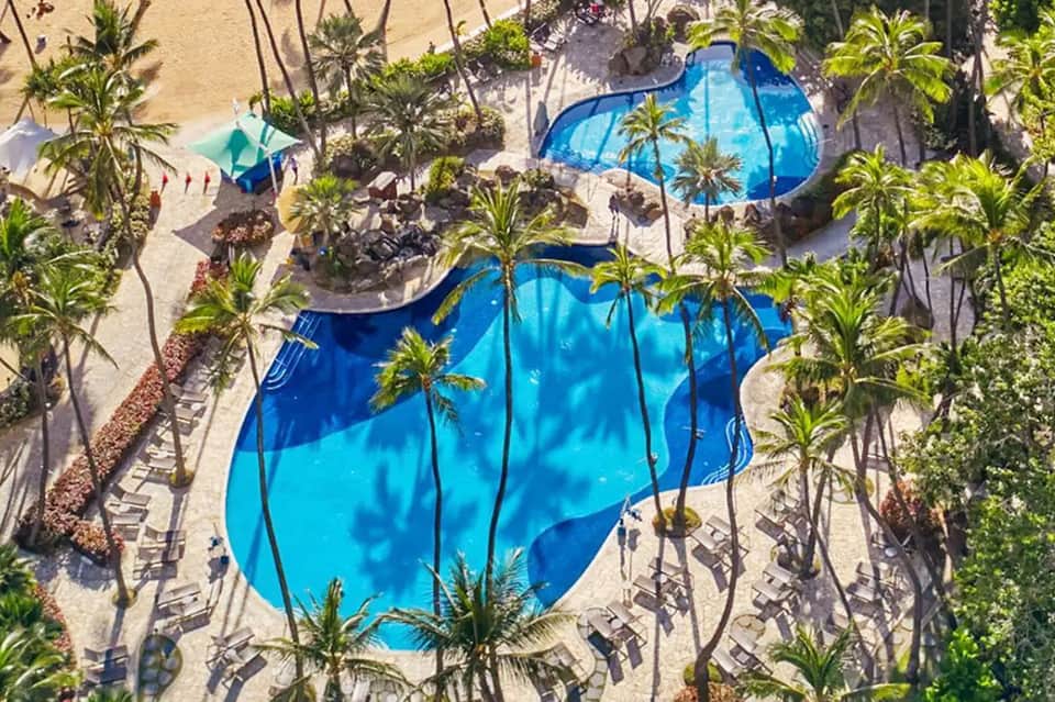 Aerial view of resort pool area with blue umbrellas, palm trees, and beach access