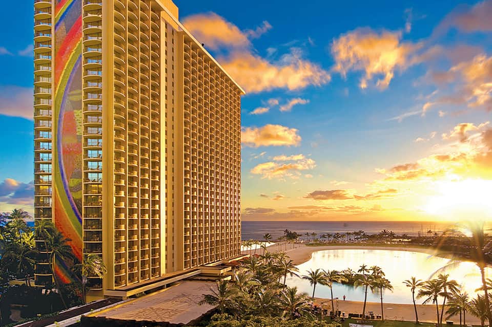 Beachfront resort tower at sunset with ocean, palm trees, and lagoon reflecting golden sky