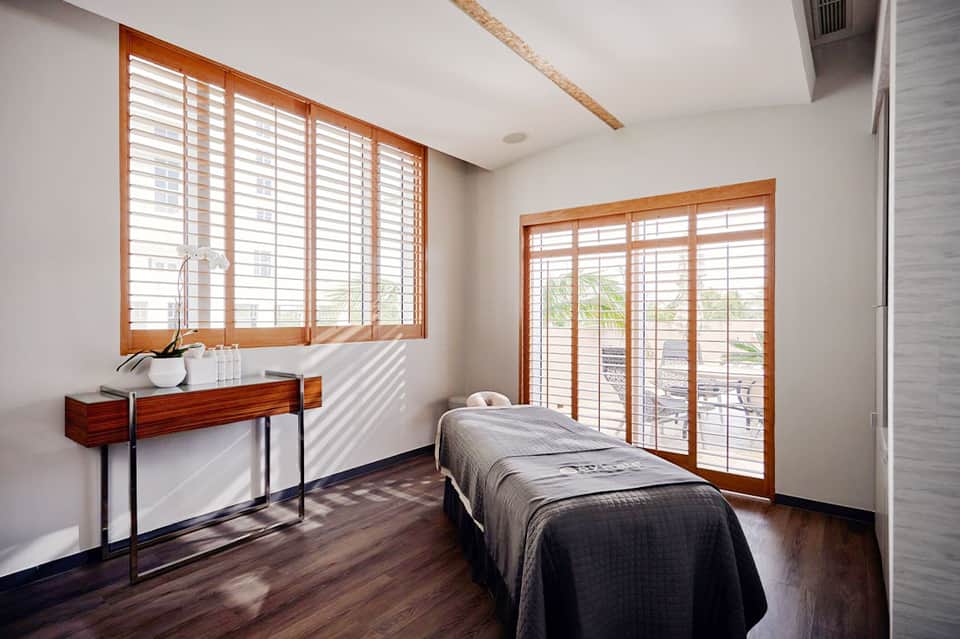 Bright spa treatment room with massage bed and ocean view through shuttered windows