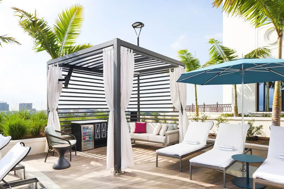 Contemporary resort deck with pergola cabana, loungers, palm trees, and waterfront views