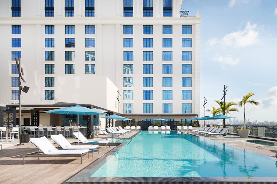 Rooftop pool with turquoise water, white lounge chairs, blue umbrellas, and high-rise building