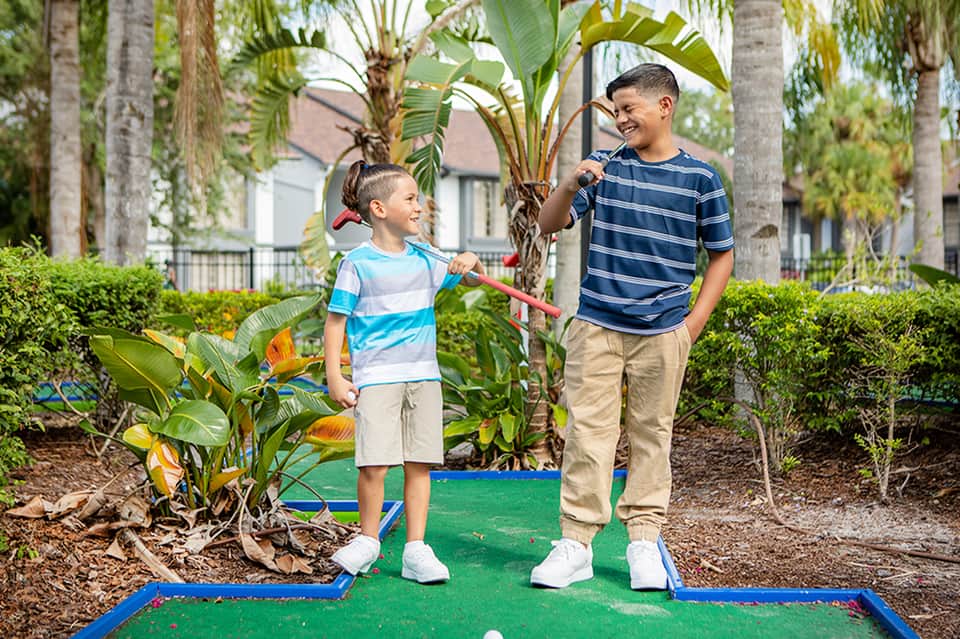 Two young boys playing mini golf outdoors surrounded by tropical plants and palm trees