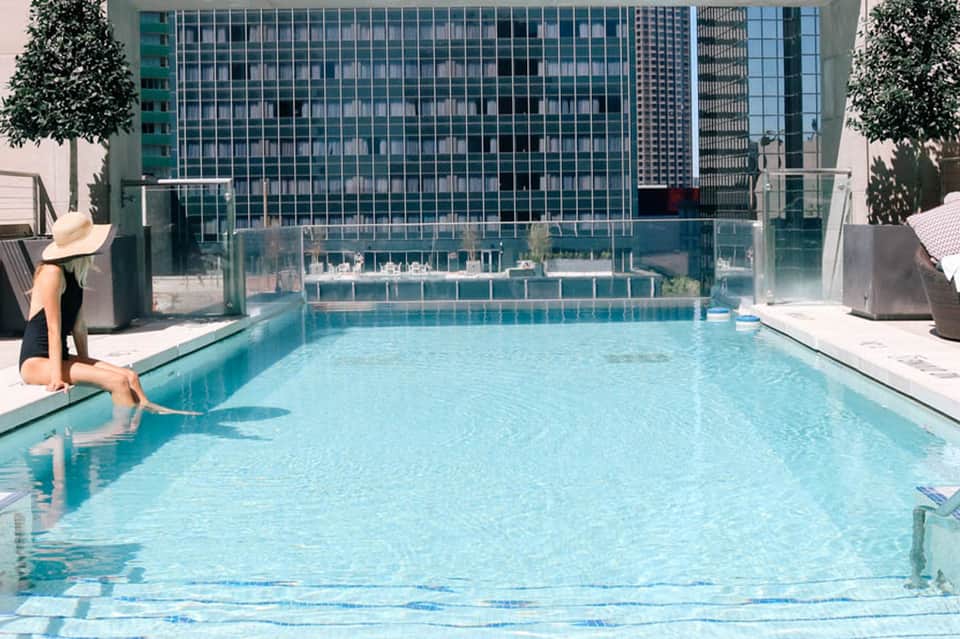Urban rooftop pool with city skyline reflection, lounge chair, and surrounding high-rise buildings