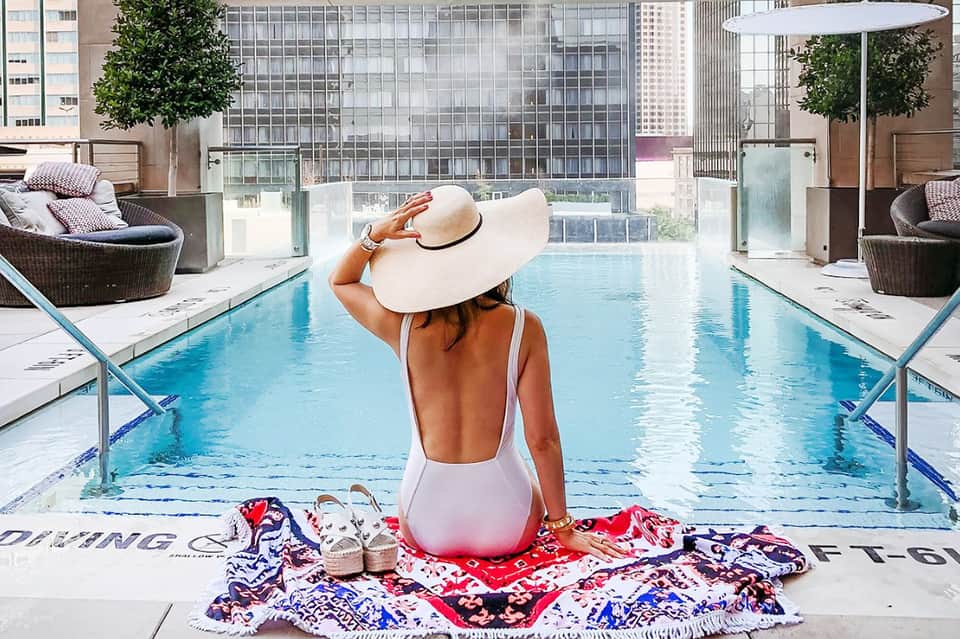 Woman in white swimsuit and hat sitting at rooftop pool overlooking city skyline