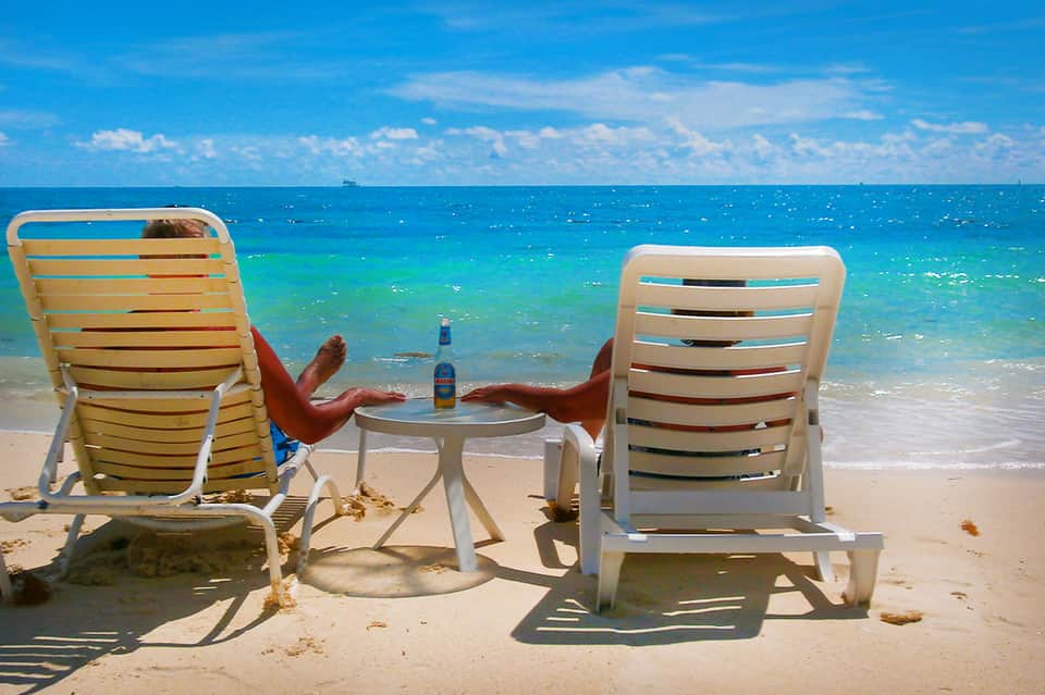 Couple relaxing in lounge chairs on pristine beach with turquoise ocean water and beer bottle