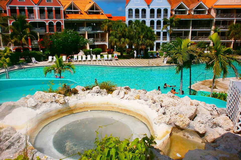 Resort pool with hot spring in foreground, colorful colonial-style buildings, and palm trees