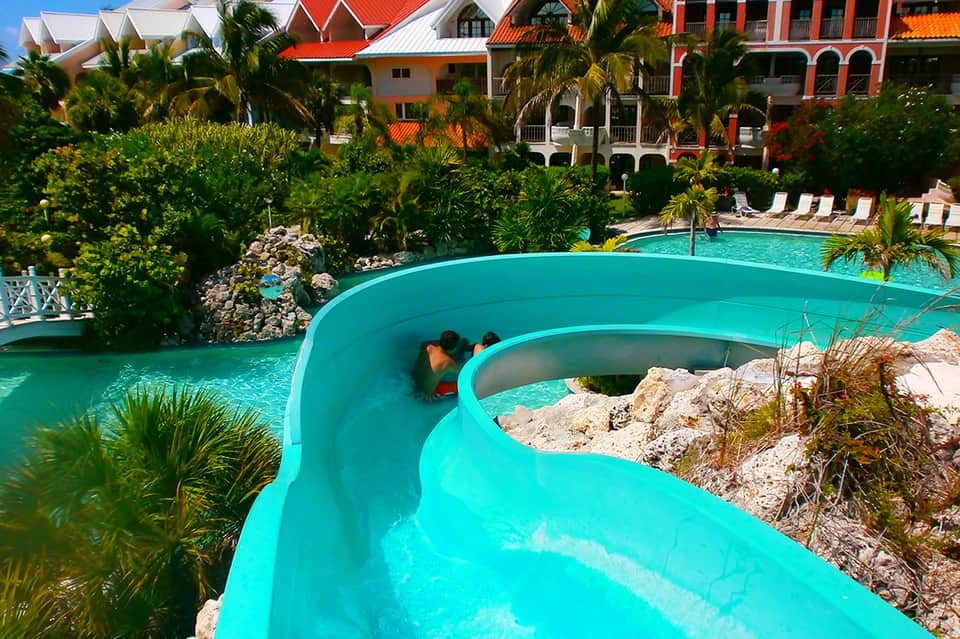 Blue water slide at resort with colorful buildings, palm trees, and lounge chairs