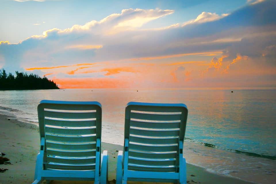 Two blue lounge chairs on a sandy beach facing a colorful sunset over calm ocean waters