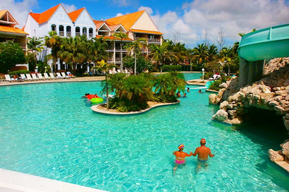 Resort pool with turquoise water, waterslide, and colonial-style orange-roofed buildings