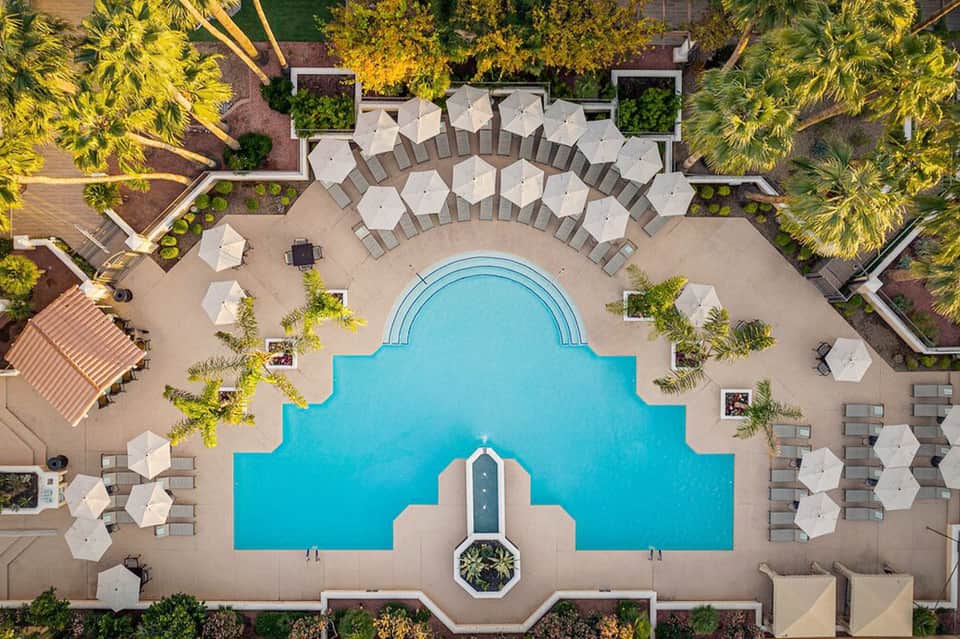 Overhead view of resort pool complex with white umbrellas, lounge chairs, palm trees, and landscaping