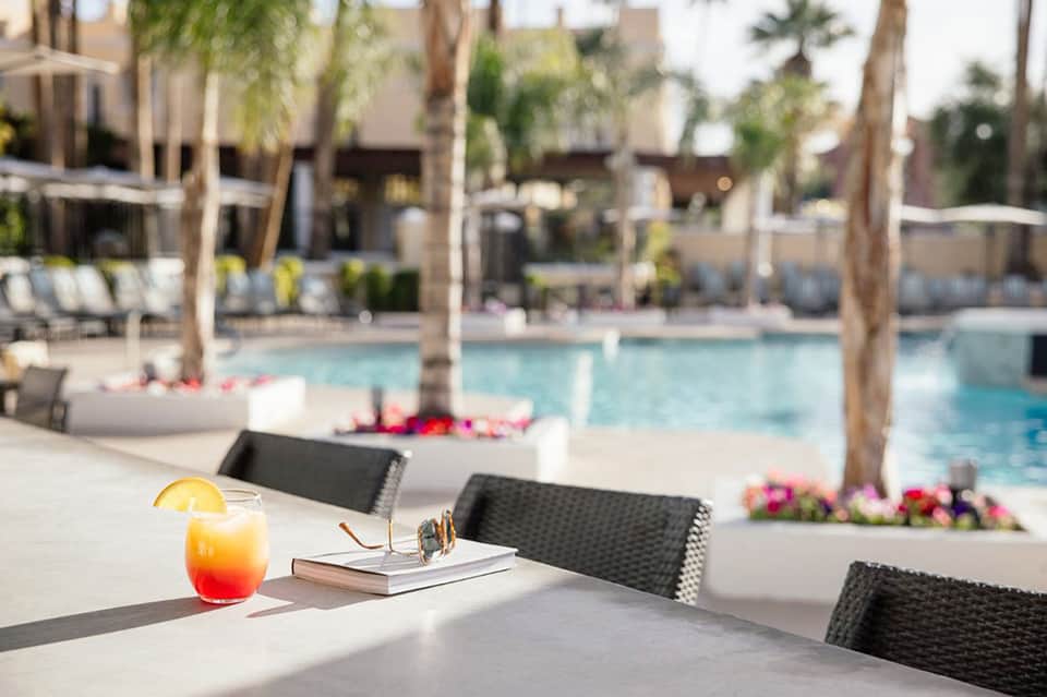 Poolside dining area with cocktail, lounge chairs, and tropical palm trees in background